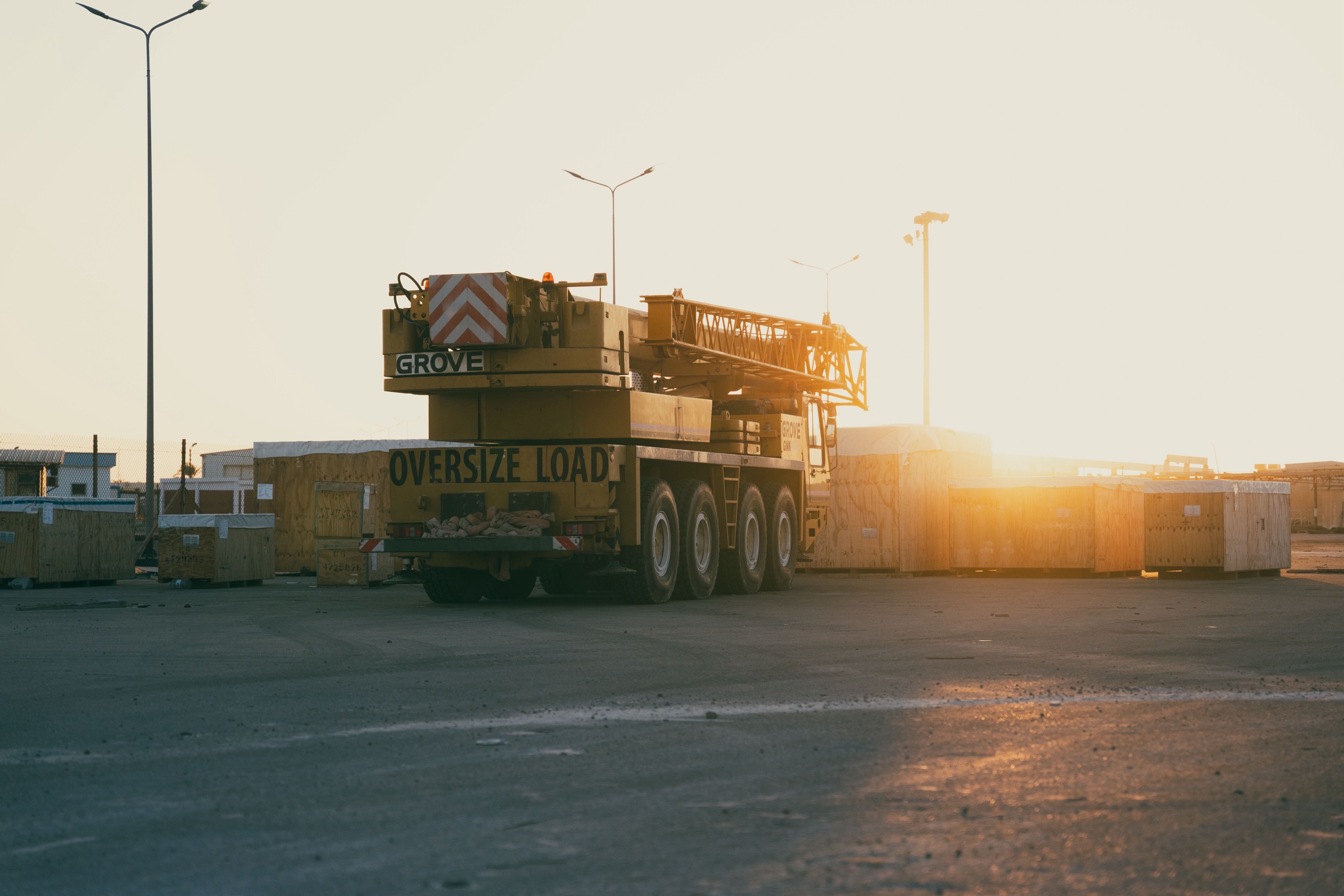 A heavy mobile crane parked in an industrial yard at dusk, low sun behind stacked timber crates