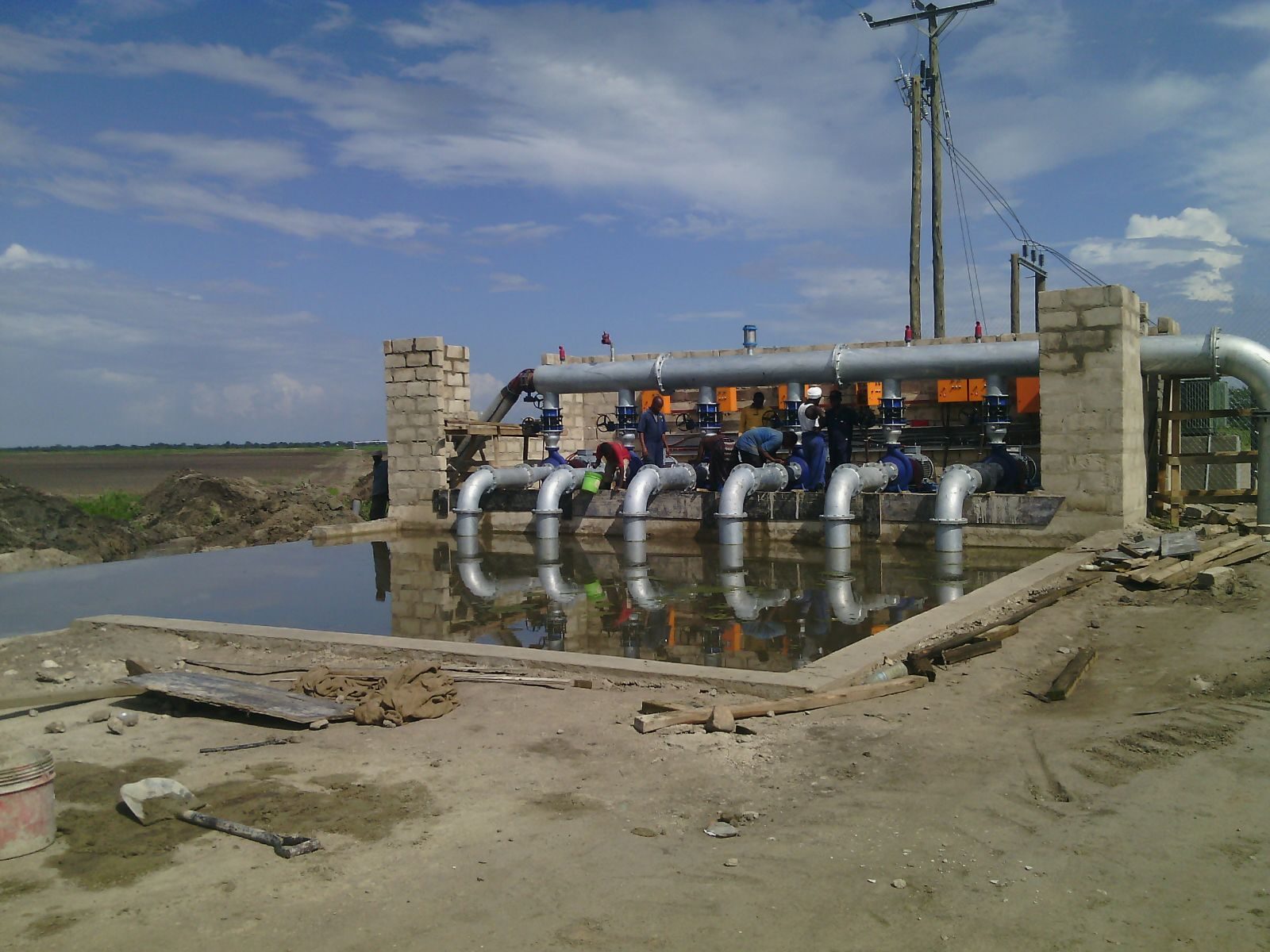 A pump station under construction beside an open water channel, with a bank of six pumps and galvanised suction pipes feeding a common delivery manifold, and workers commissioning the units