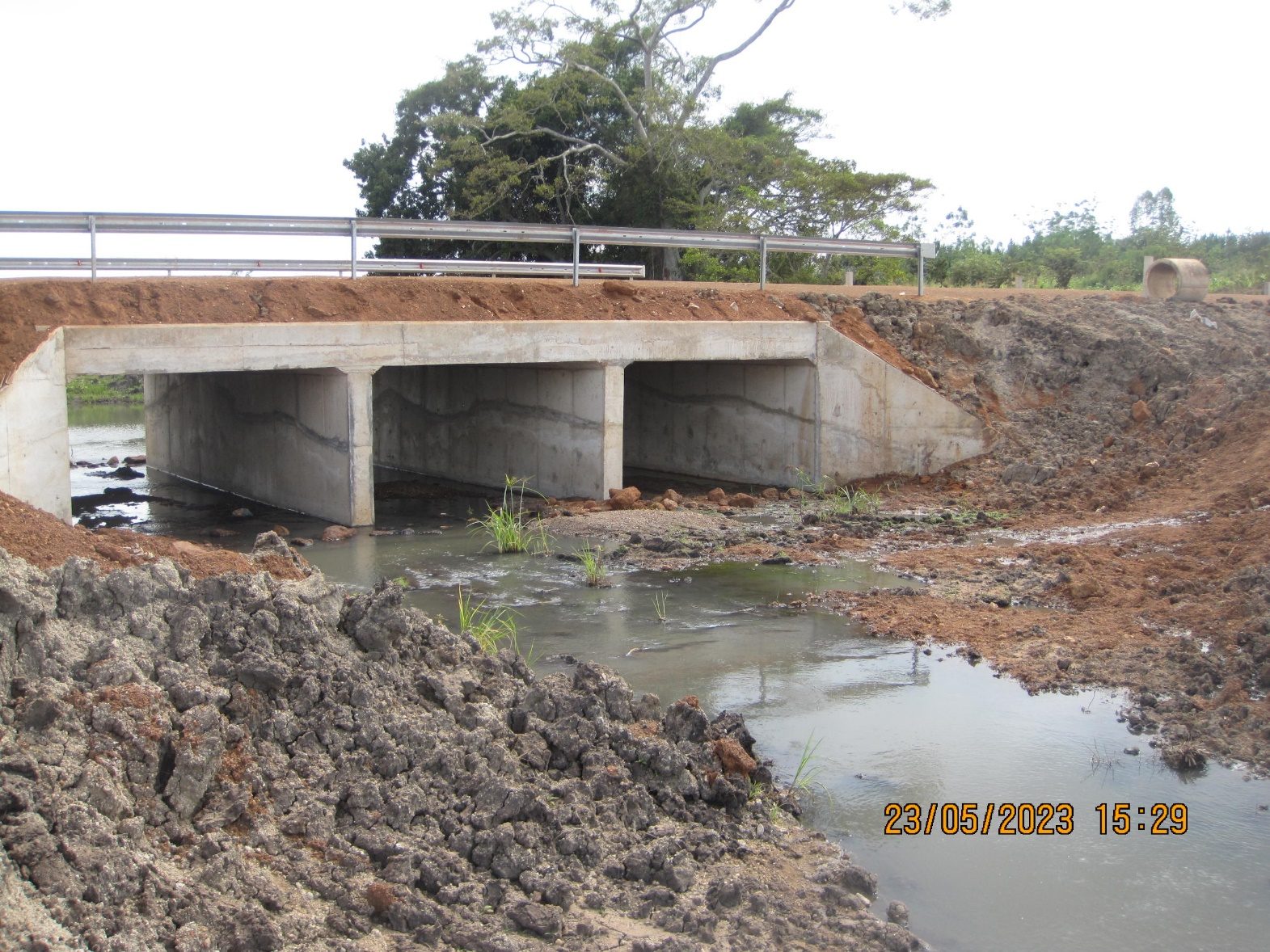 A completed three-cell reinforced concrete box culvert carrying a gravel road with steel guardrails over a stream