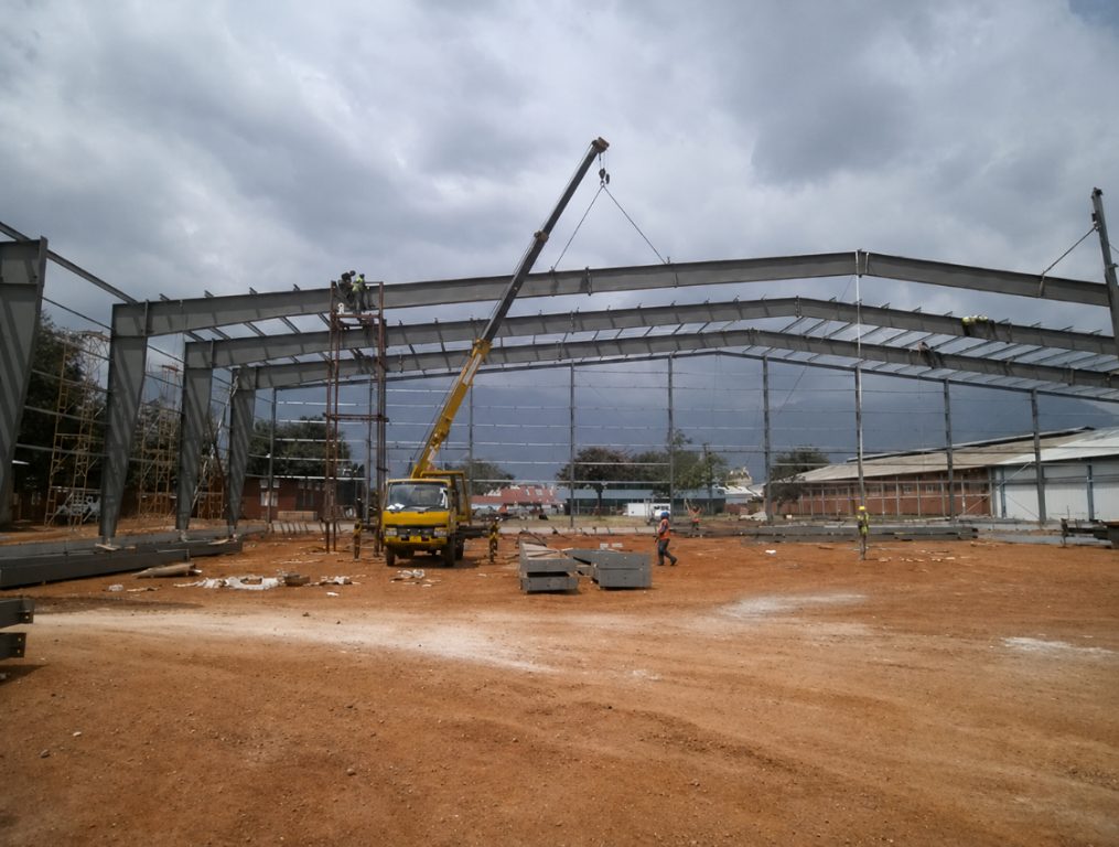 A steel portal-frame building under erection, with a mobile crane lifting a rafter into place and workers on ladders securing steelwork