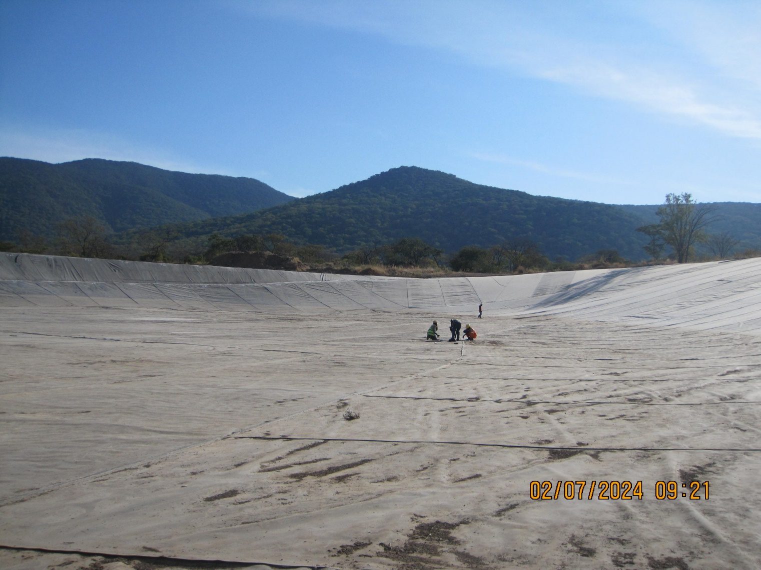 A wide tailings storage facility basin fully lined with geomembrane, with three workers kneeling on the liner in the middle distance and forested hills on the horizon