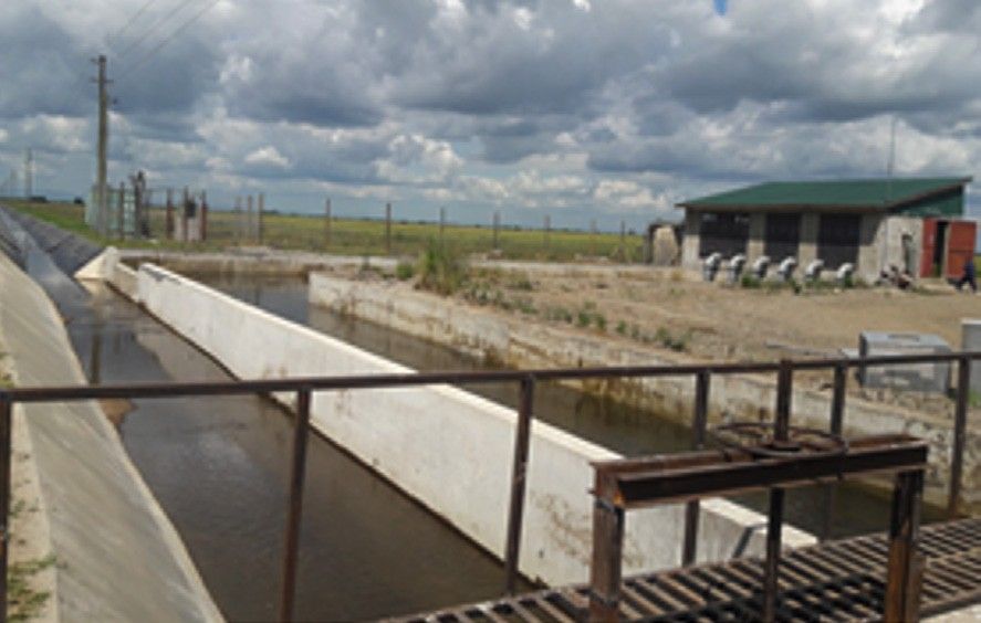 A concrete-lined canal with a steel footbridge in the foreground, running towards a green-roofed pump house on open plains