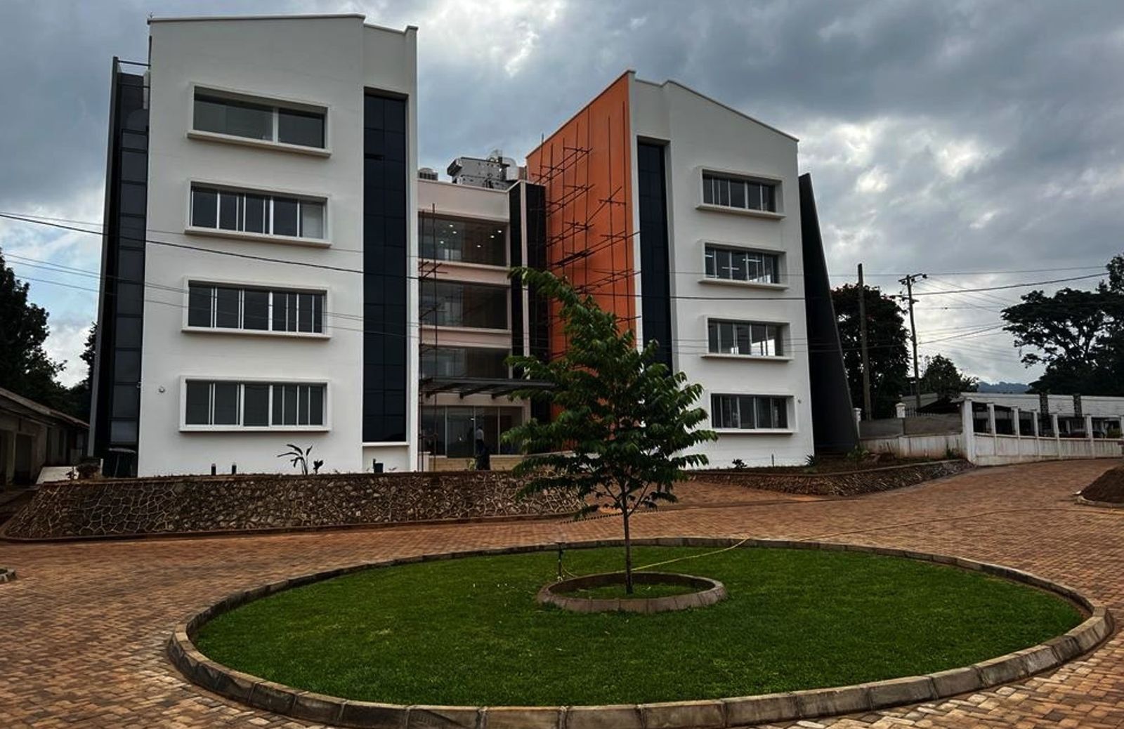 A completed four-storey laboratory building in white render with dark glazing and an orange feature panel, fronted by a paved forecourt and a circular lawn with a young tree