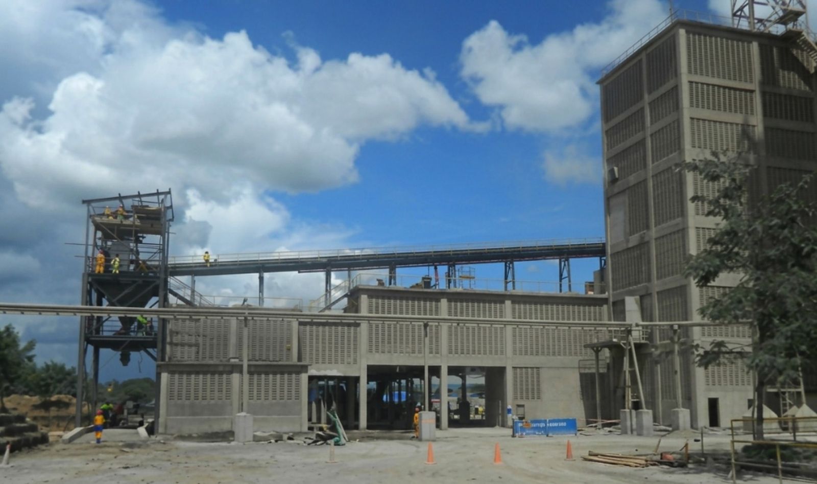 A tall concrete loading tower beside a steel gantry and conveyor bridge at a cement factory, with workers in high-visibility vests at the base