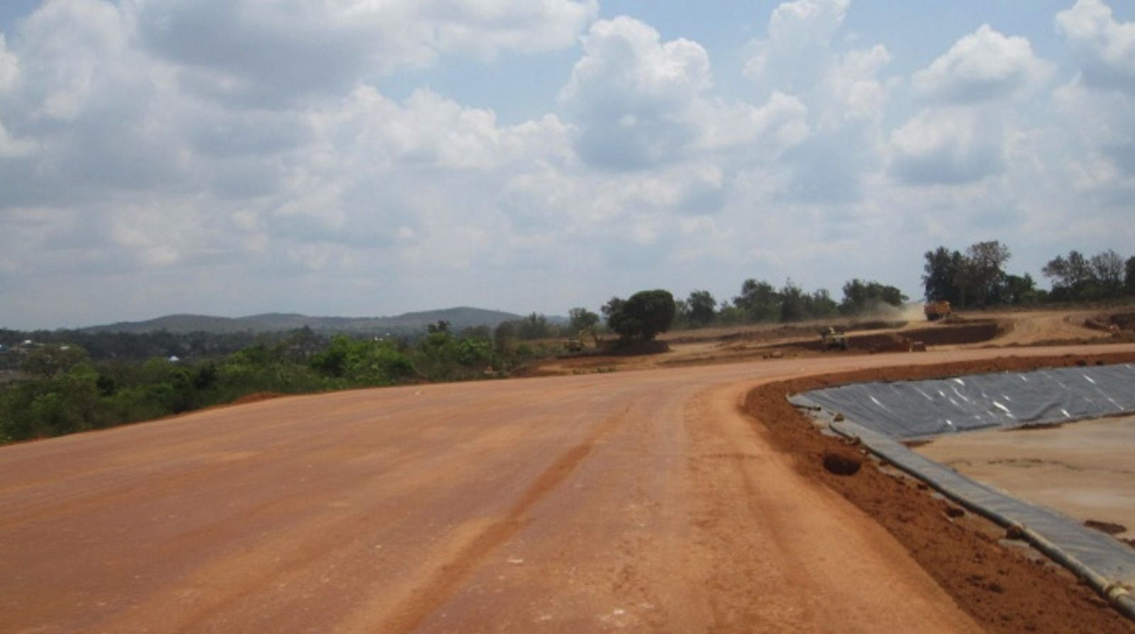 A wide compacted earth access road curving around the edge of a geomembrane-lined cell