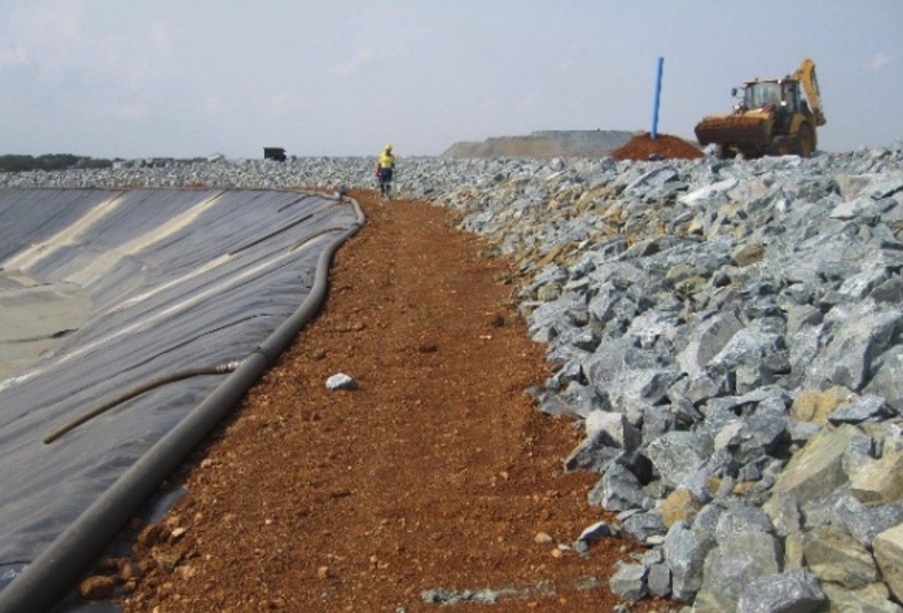 A rockfill embankment beside a geomembrane-lined slope, with a backhoe loader working on the crest