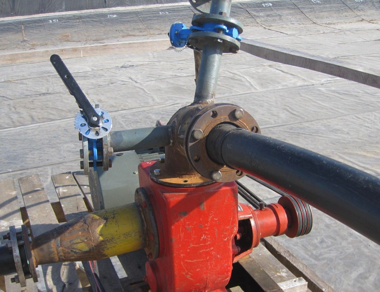 A red pump and valve assembly connected to black pipework, mounted on timber on a geomembrane-lined basin floor