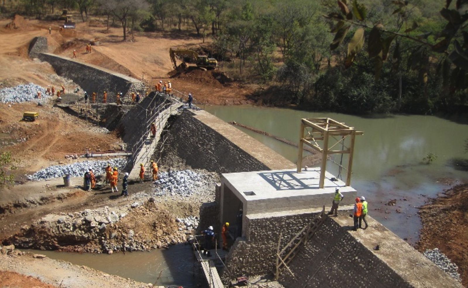 A masonry water dam under construction, with a large crew in orange overalls working along the sloping masonry face, a concrete outlet structure and the impounded water beyond