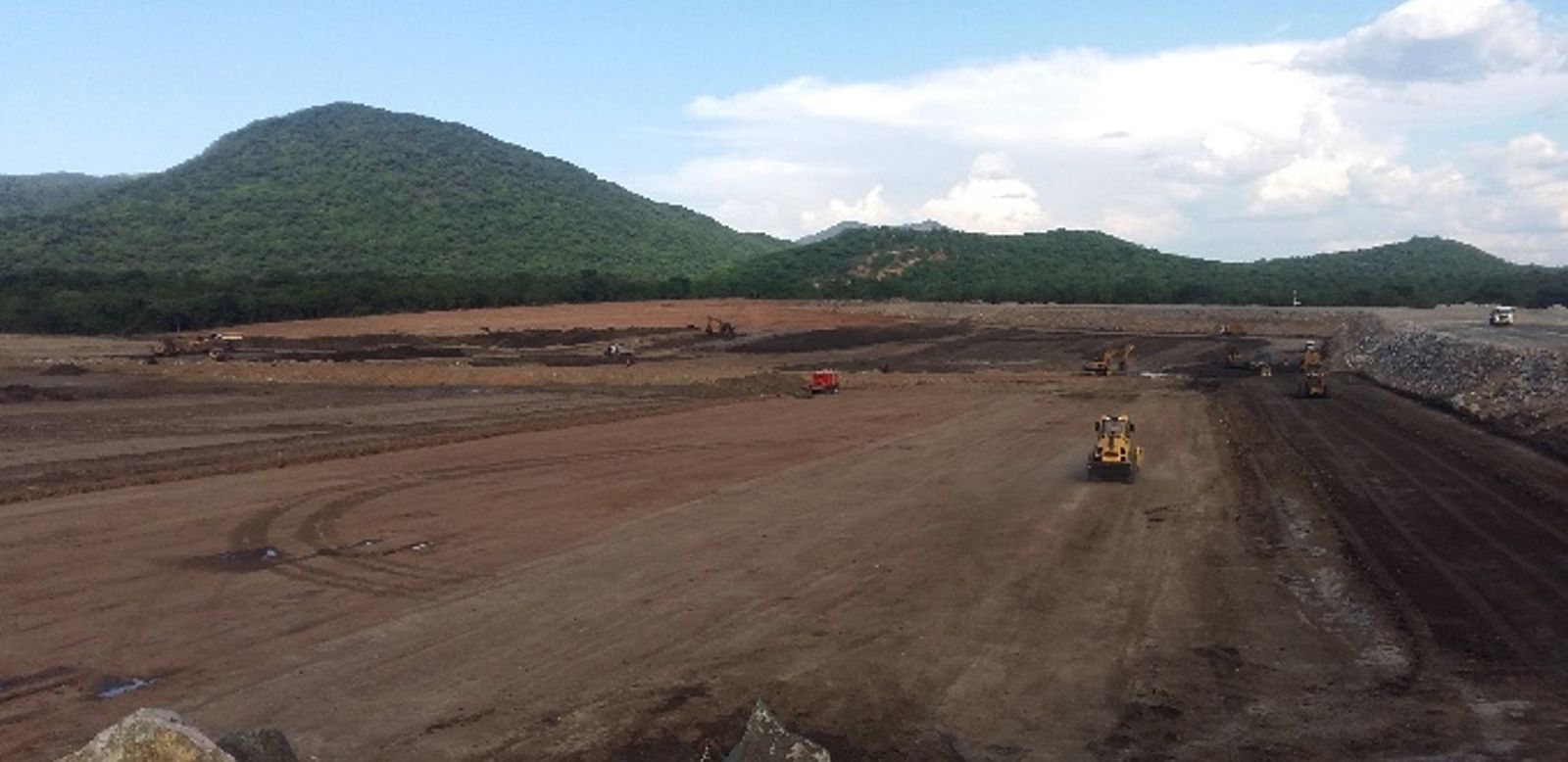 A wide basin being shaped by earthmoving machinery at the foot of a forested mountain