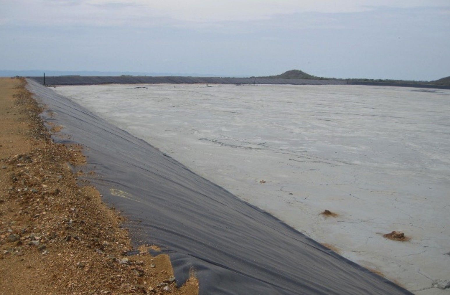 A tailings basin filled with pale grey tailings, bounded by a geomembrane-lined embankment slope
