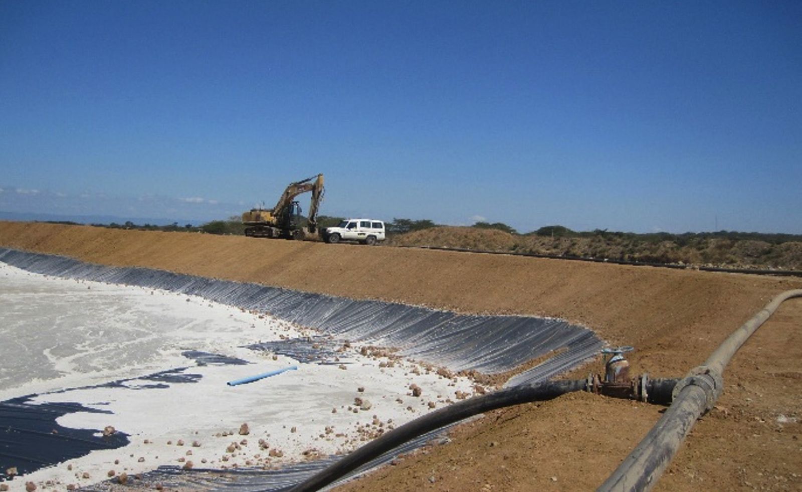 A geomembrane-lined tailings cell with a pipeline along the embankment crest and an excavator and a vehicle working beyond
