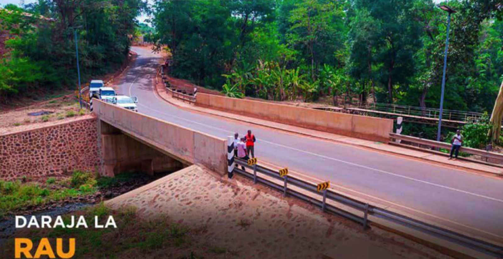 A completed road bridge with concrete parapets and steel guardrails carrying a sealed road through dense green vegetation, overlaid with the words Daraja la Rau