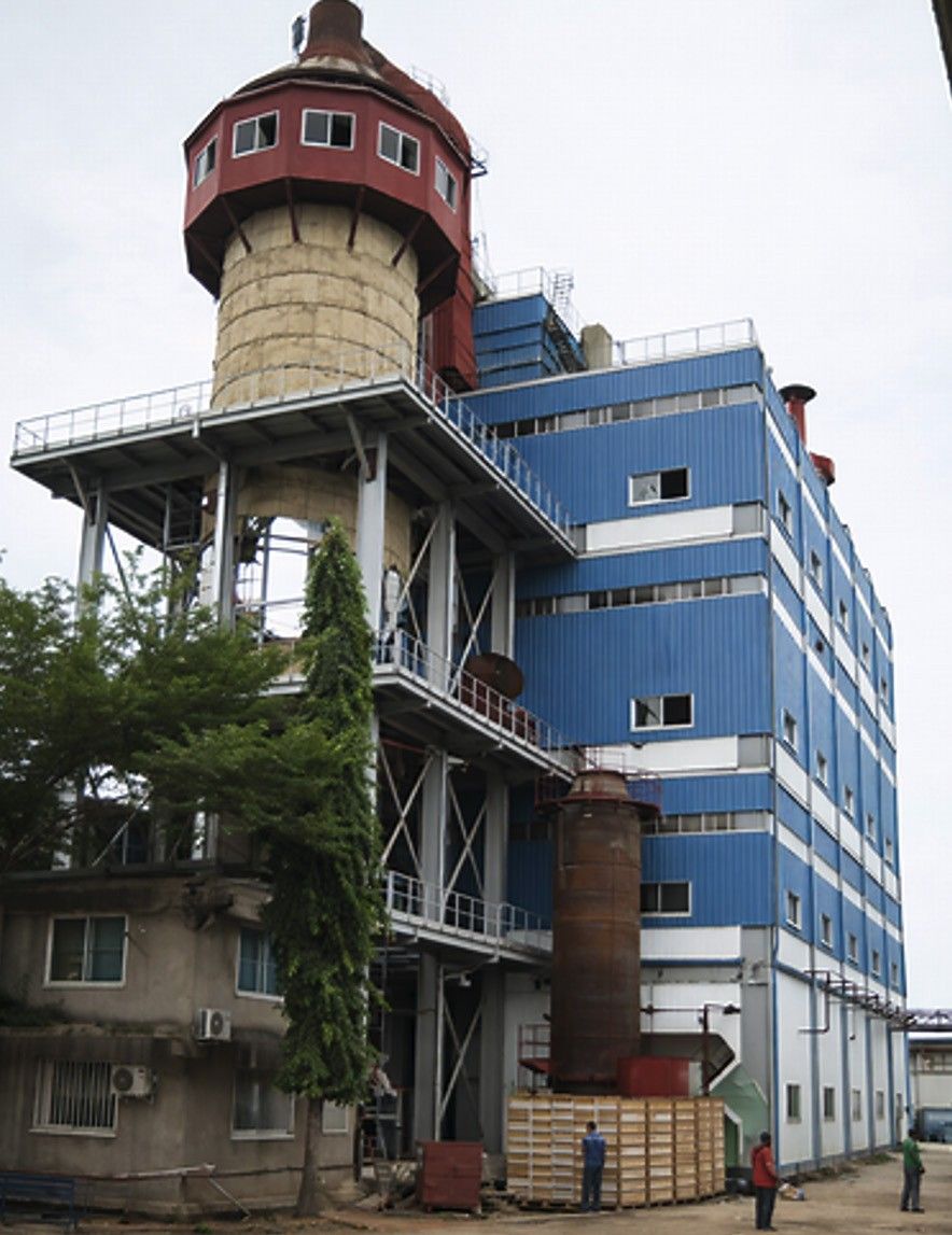A multi-storey industrial building clad in blue panels beside a tall cylindrical tower with a red-roofed top, with workers and timber crates at its base