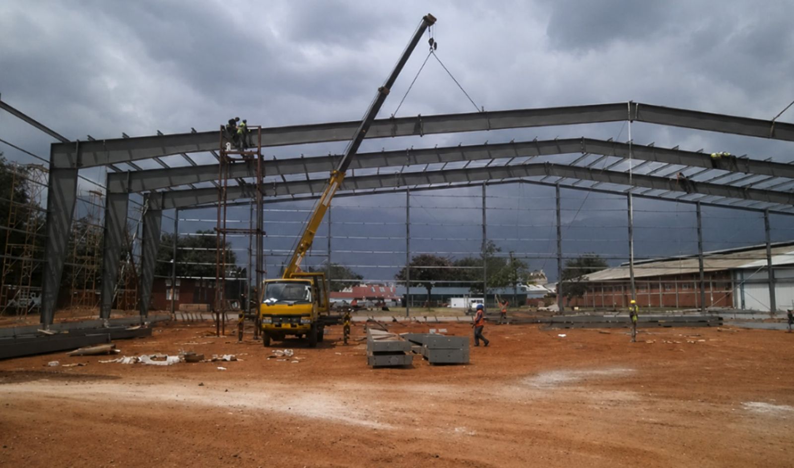 A steel portal-frame building under erection, with a mobile crane lifting a rafter into place and workers on scaffolding securing the steelwork