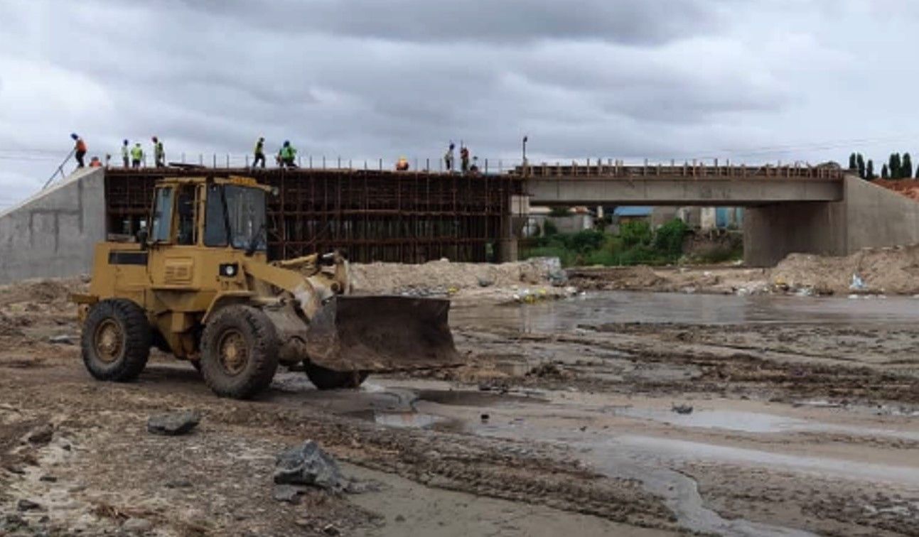 A bridge under construction over a river bed, with formwork and reinforcement on the deck, workers along the top and a wheel loader working in the foreground